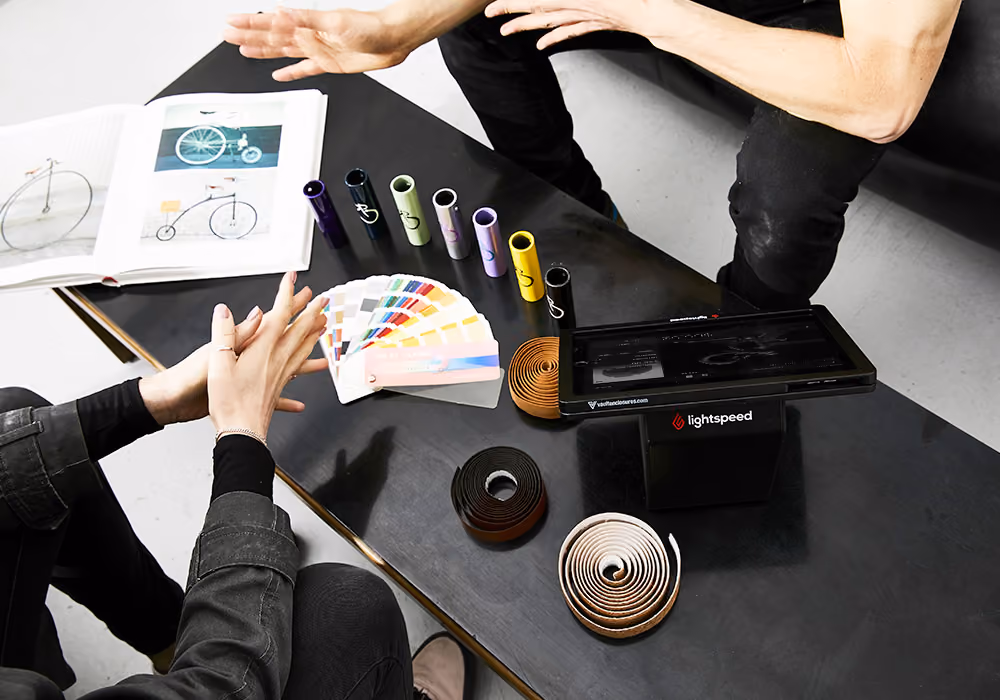 Two people seated at a table with bike handlebar tapes, a color palette, and an open book with bicycle images.