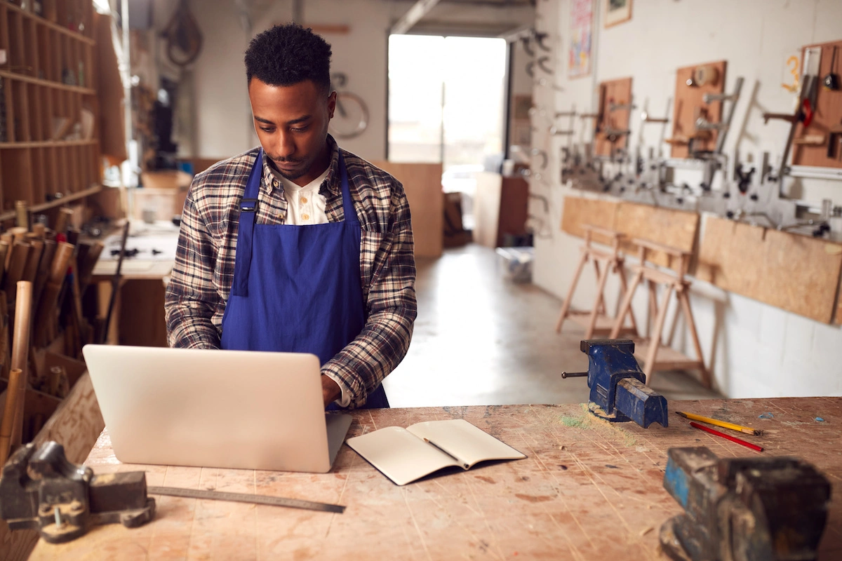 Man in a blue apron working on a laptop in a woodworking shop with tools and notebooks nearby.