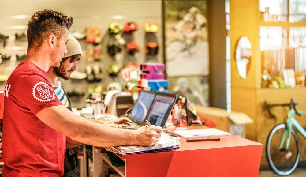 Two men working at a shop counter with laptops, helmets, and bicycles in the background.
