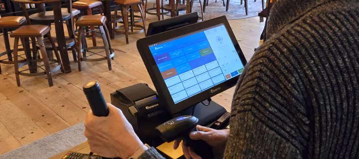 Person using touchscreen POS system with attached receipt printer in a restaurant with wooden chairs.