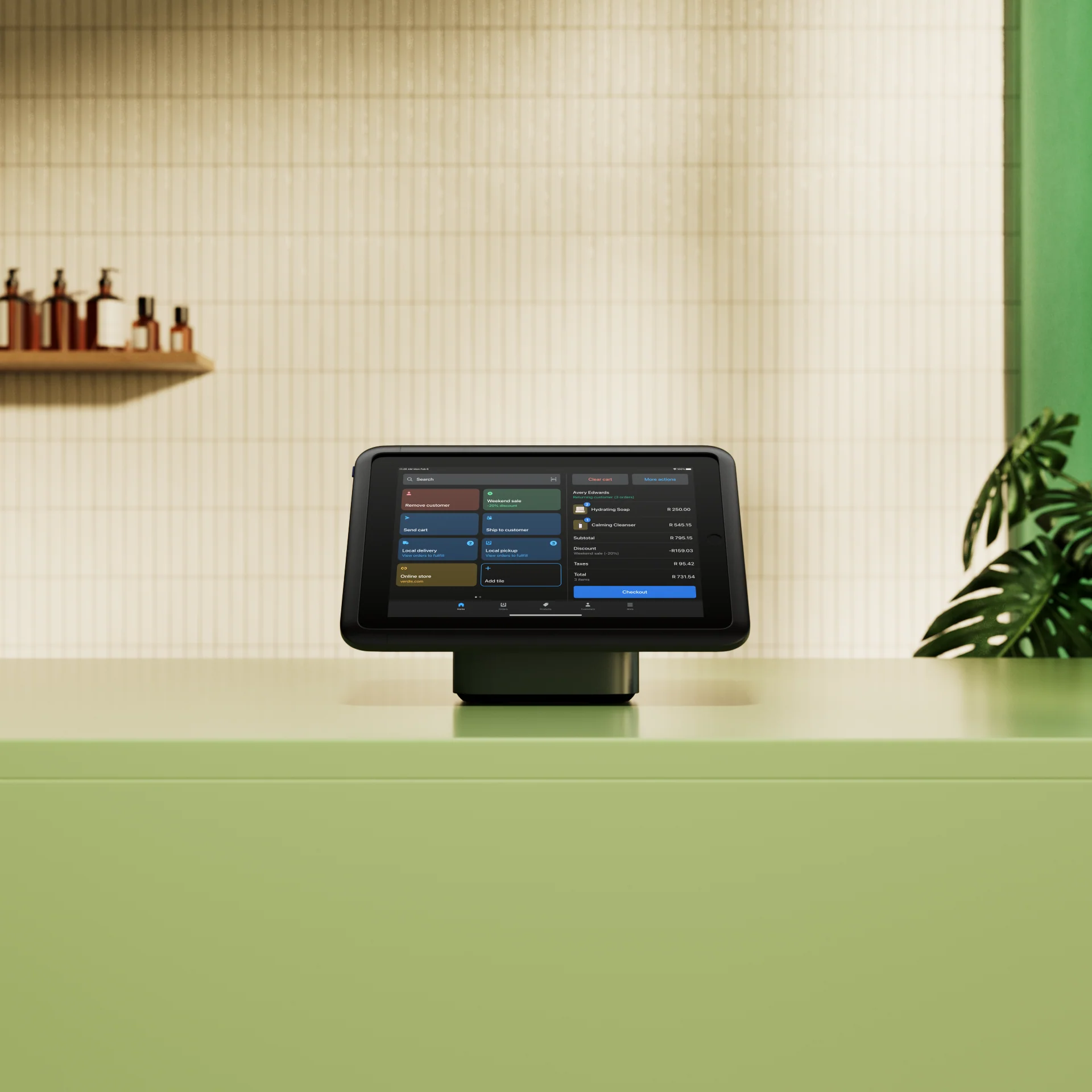 Touchscreen point-of-sale device on green counter with tiled wall and shelf holding bottles in background.