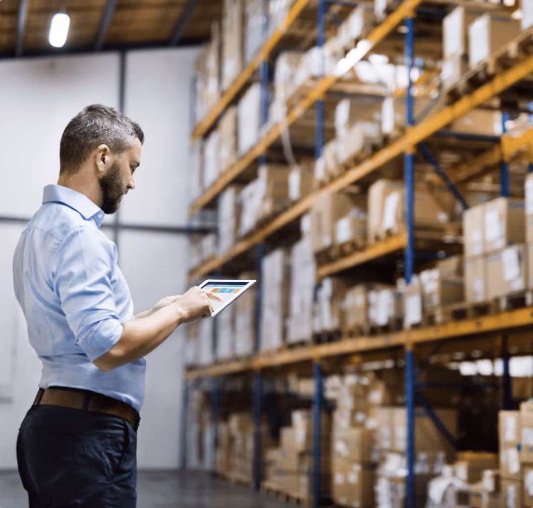 Man using a tablet in a warehouse aisle with shelves full of stacked cardboard boxes.