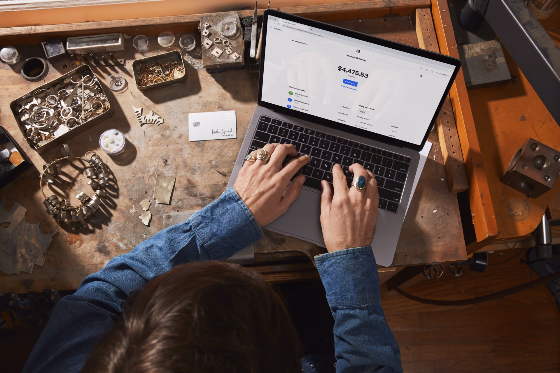 Person with rings typing on laptop showing $4,475.53 balance on cluttered wooden workbench.