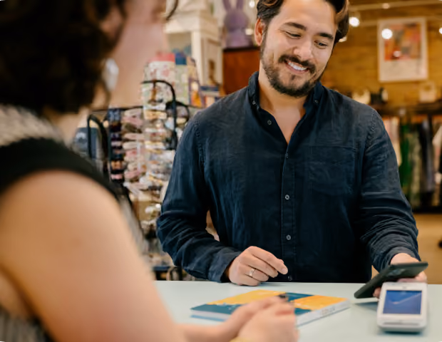 Smiling man paying with smartphone at a store counter while woman watches and waits.