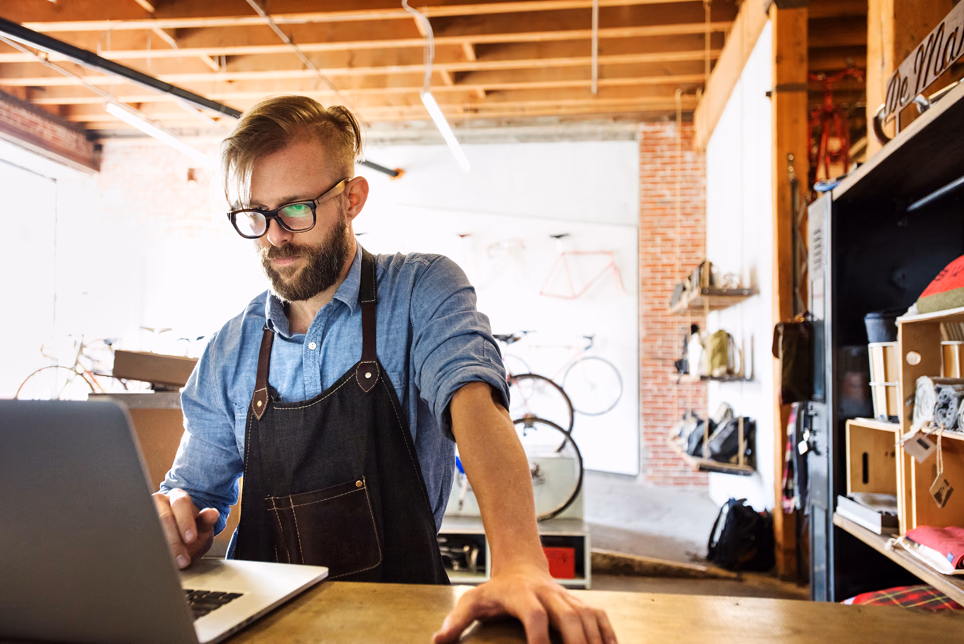 Man with glasses and apron using a laptop in a bicycle repair shop with bikes and gear in the background.