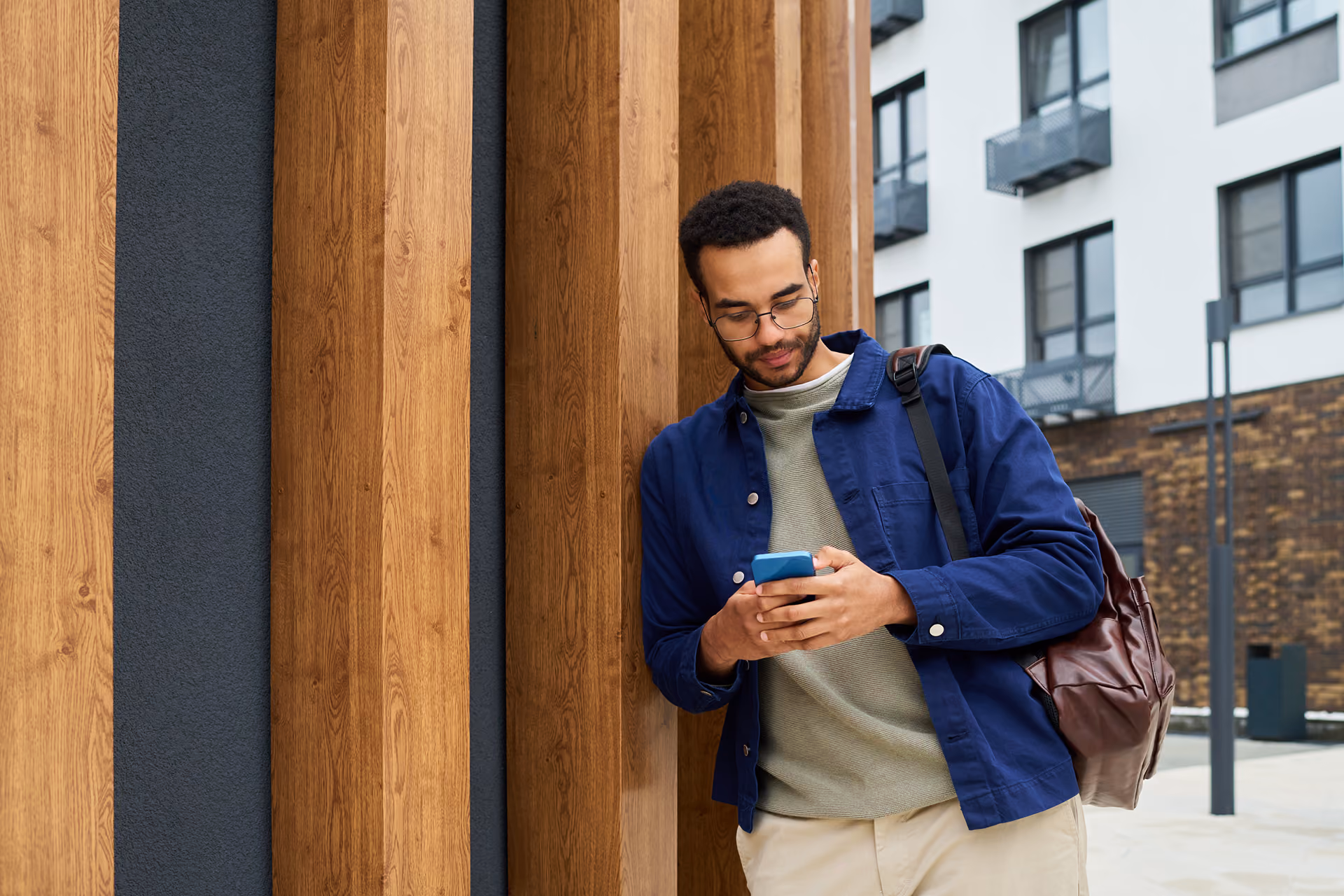 Man wearing glasses and blue jacket leaning against wooden pillars while using a smartphone outdoors.