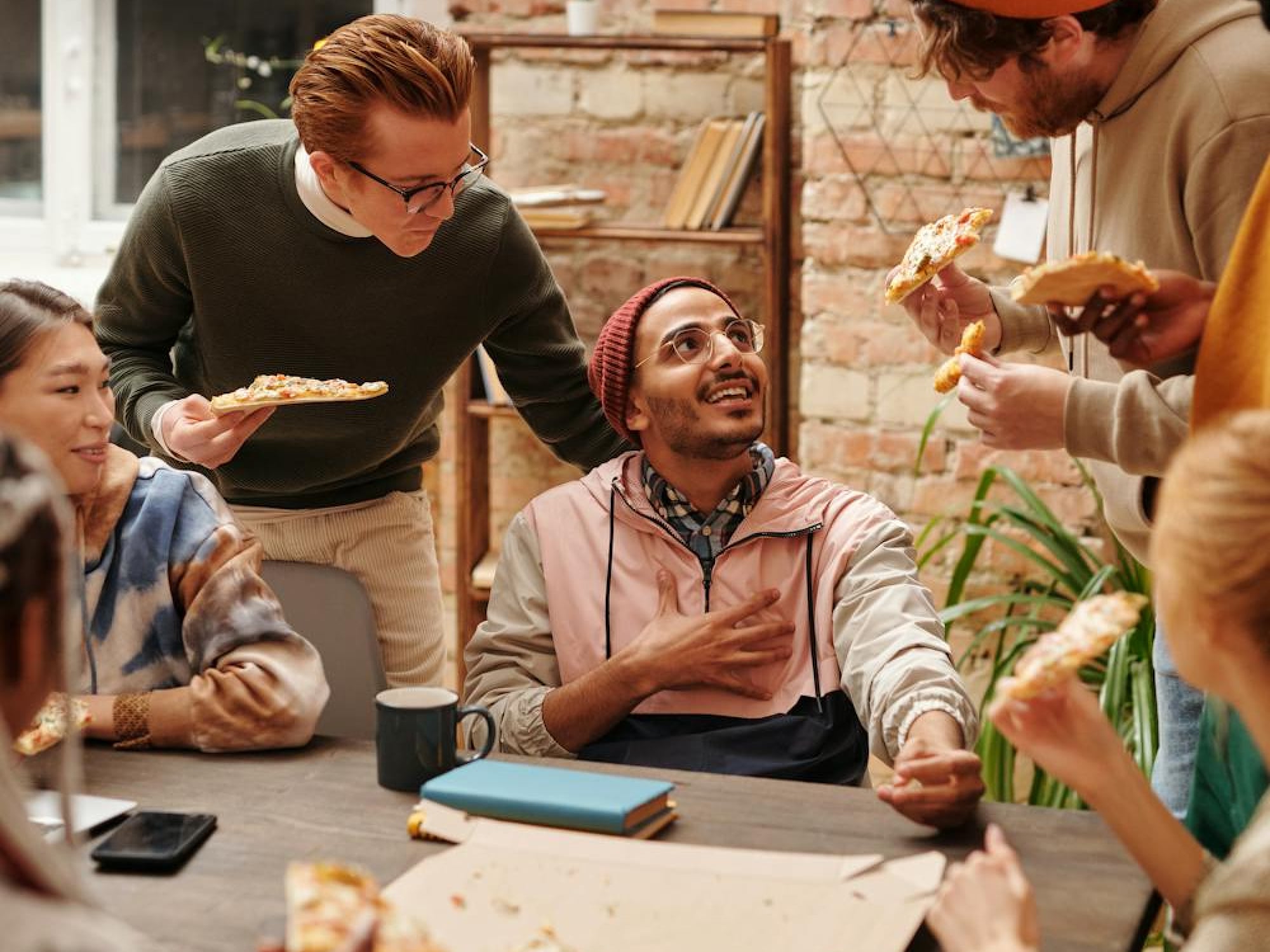 Group of young adults enjoying pizza together indoors, engaged in lively conversation around a wooden table with pizza slices, books, a mug, and a smartphone visible.