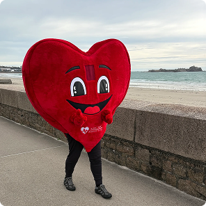 Person walking on a sidewalk near the beach wearing a large red heart costume with a happy face and thumbs up, with ocean and cloudy sky in the background.