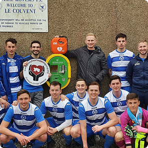 Group of young men posing in blue and white soccer uniforms in front of a wall with a sign reading 'Le Couvent' and a defibrillator mounted on it.