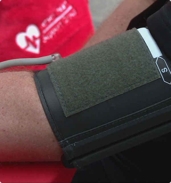 Close-up of a person’s arm with a blood pressure cuff wrapped around it, connected to a tubing, against a blurred background showing a red fabric with a white heart and medical symbol.