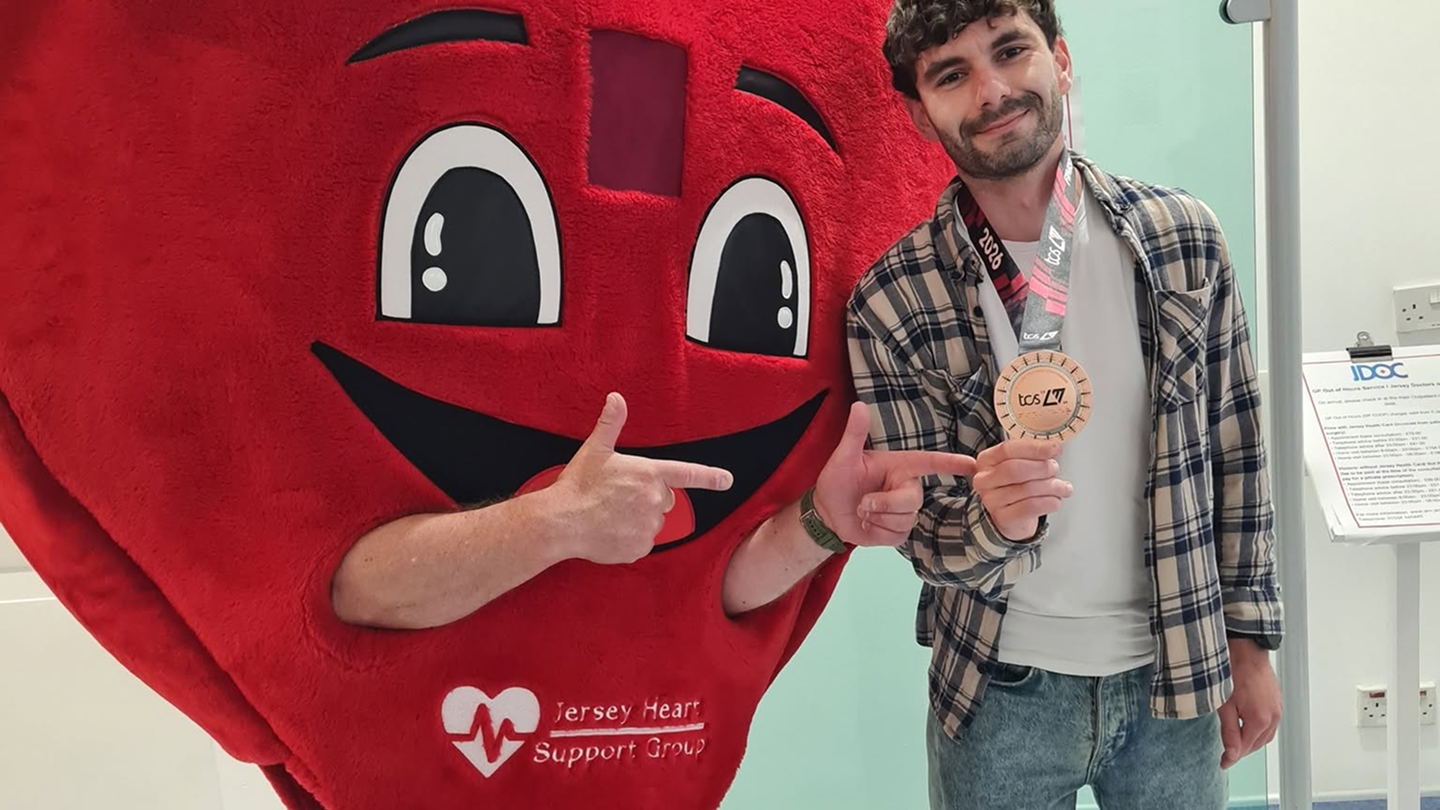 A man in a plaid shirt and jeans stands next to a person in a large red heart costume with a face, both pointing at a circular medal the man is holding, which has a 'tcs' logo and the year 2026 on the ribbon.