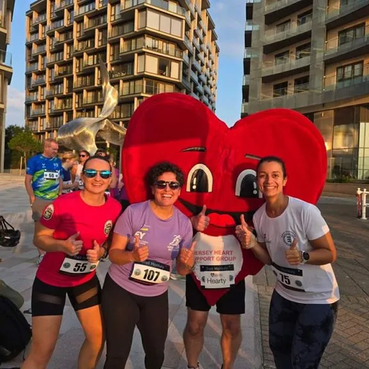 Three women with race bibs smiling and giving thumbs up pose with a person in a large red heart mascot costume in an urban outdoor setting with modern buildings in the background.