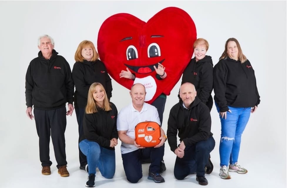 Group of eight people posing in a studio with a white background including one person in a large red heart mascot costume. Most people are wearing black hoodies with a small red heart logo on them. A man in the center kneels holding an orange AED (automated external defibrillator) device.