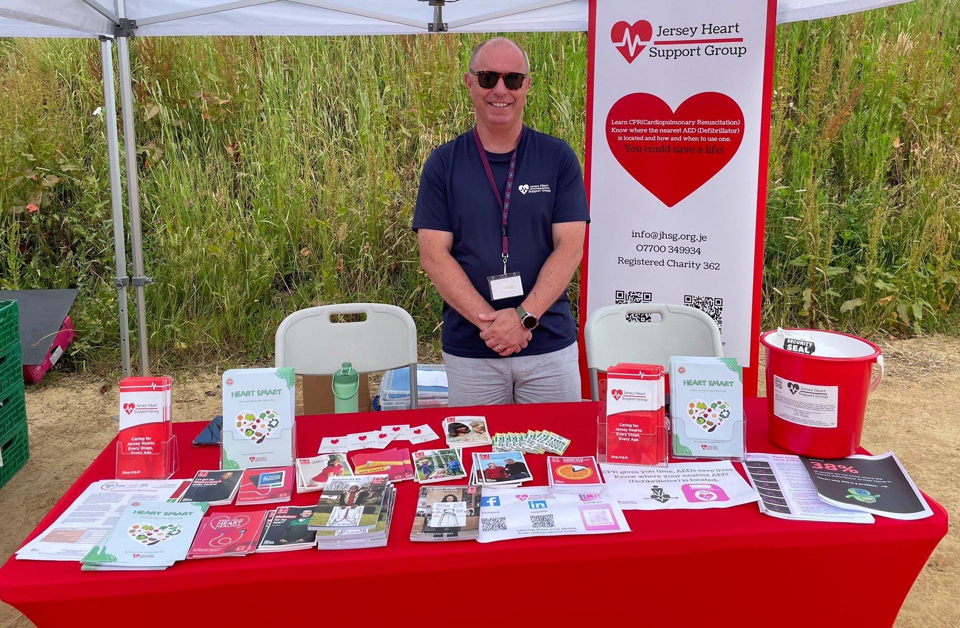 Man wearing sunglasses and a Jersey Heart Support Group t-shirt stands behind a table covered with informational brochures, flyers, and booklets about heart health, CPR, and AED use at an outdoor event under a white canopy, with a Jersey Heart Support Group banner behind him.