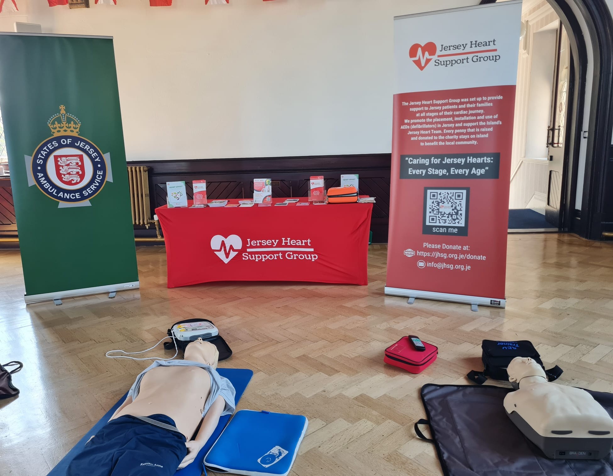 Indoor scene showing a Jersey Heart Support Group table with brochures and AED device, flanked by States of Jersey Ambulance Service and Jersey Heart Support Group banners, with two CPR training mannequins lying on mats on the wooden floor, alongside AED trainers and related equipment.