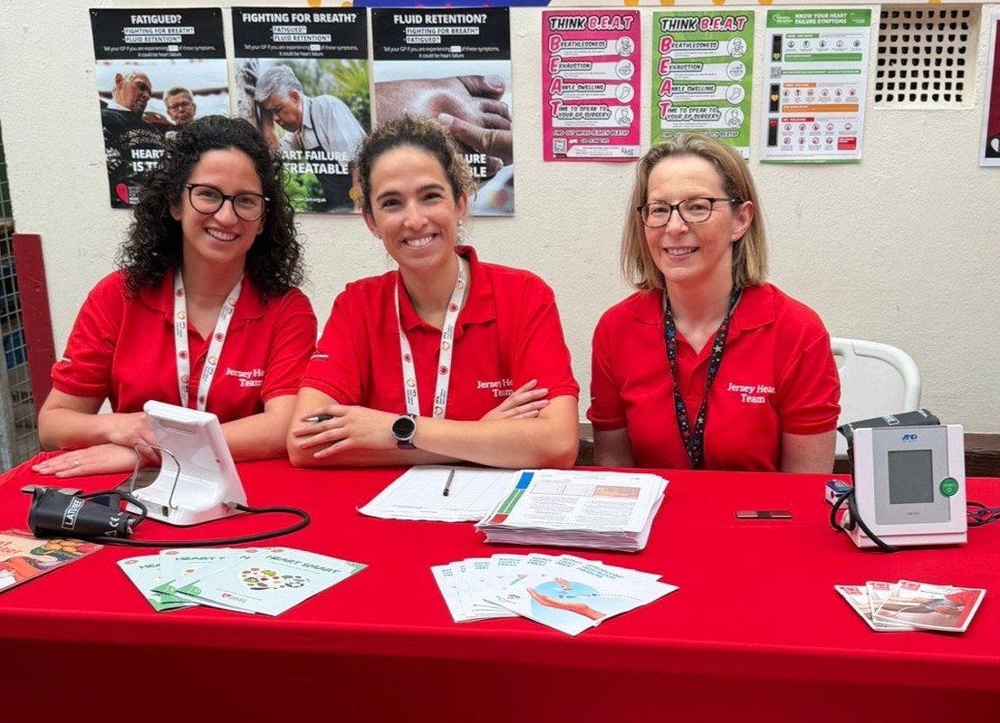 Three women wearing red Jersey Heart Team polo shirts sit behind a table with red cloth, displaying health pamphlets, paperwork, and medical devices for blood pressure and heart monitoring, against a wall with health awareness posters about symptoms and heart failure.