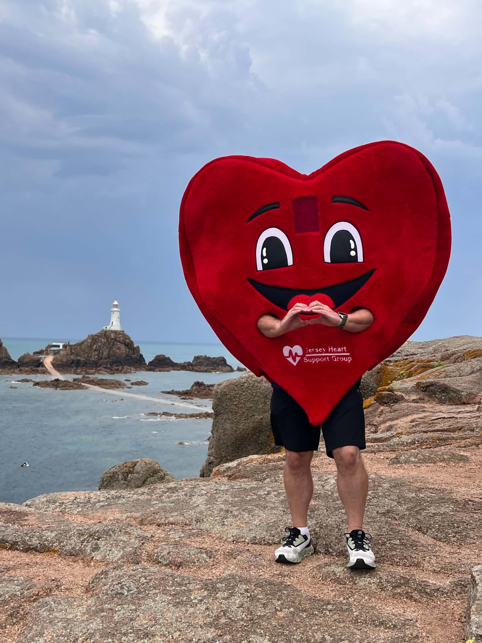 Person standing on rocky terrain near the sea wearing a large red heart costume with a smiling face and the words 'Jersey Heart Support Group' printed on it, making a heart shape with their hands, with a lighthouse on a rocky island in the background under a cloudy sky.