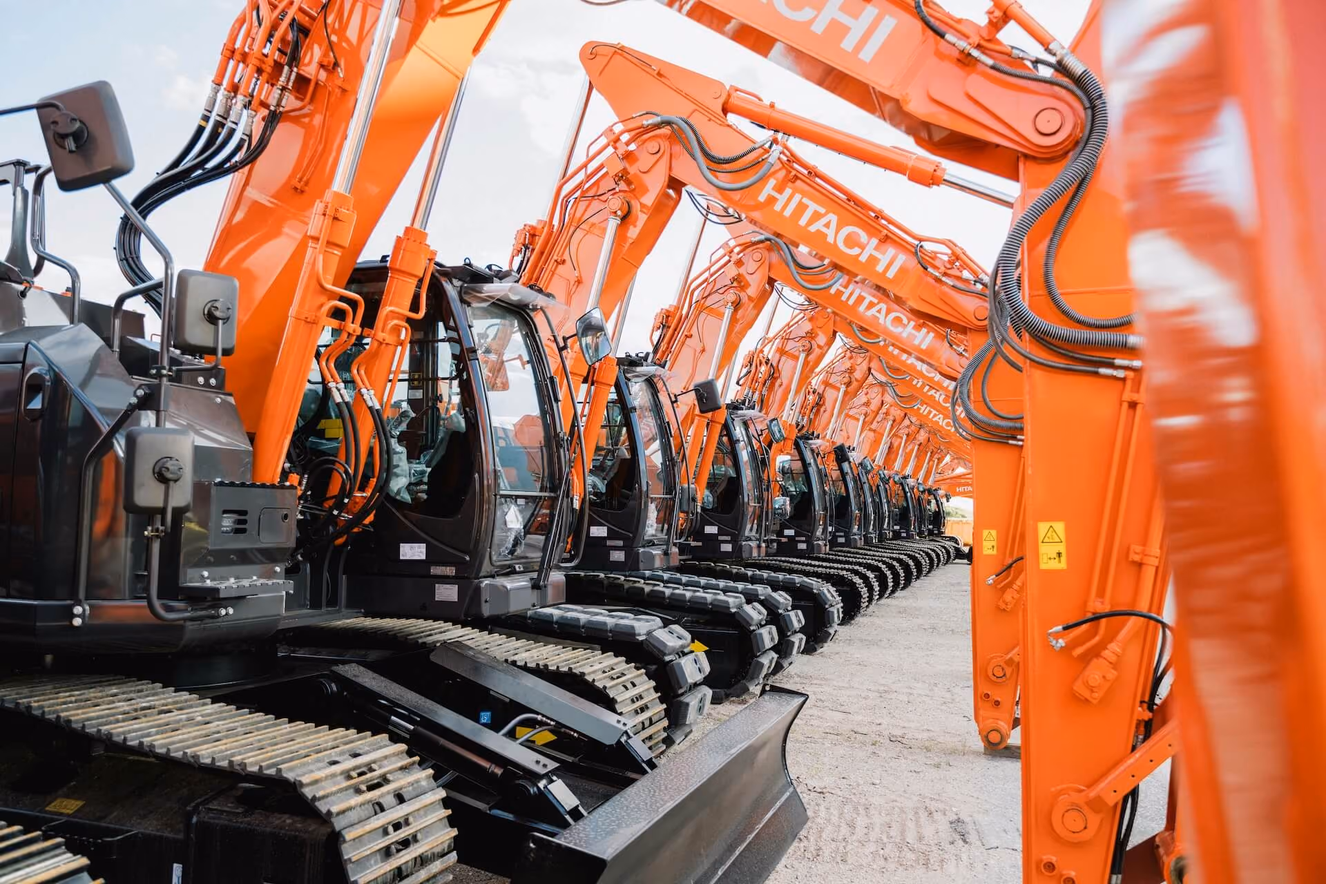 A row of large orange and black Hitachi excavators lined up outdoors on a concrete surface, viewed from the side showing their tracks and hydraulic arms.