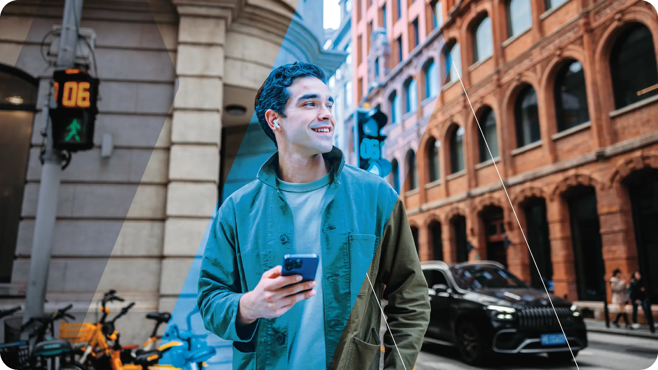 Jeune homme souriant, tenant un smartphone avec des écouteurs sans fil, marchant dans une rue urbaine avec des bâtiments en briques et des vélos garés, près d'un feu piéton vert.