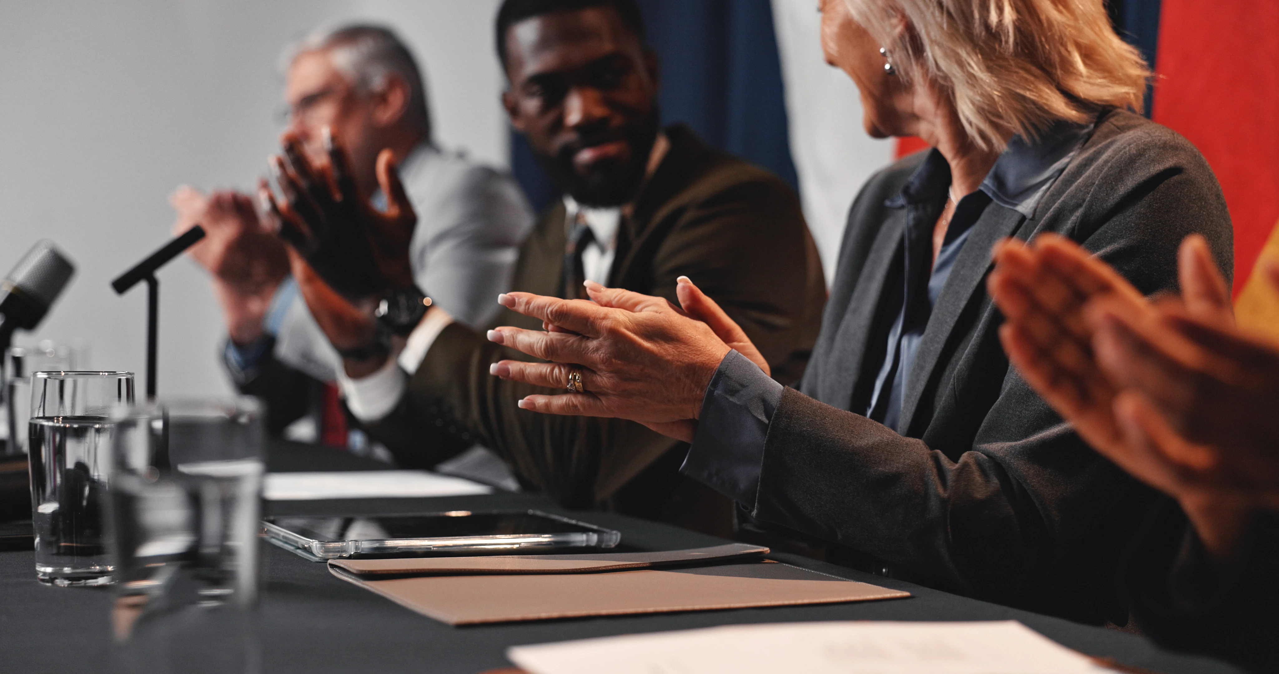 A diverse group of professionals sitting at a table clapping, with microphones and glasses of water in front of them, suggesting a formal meeting or panel discussion.