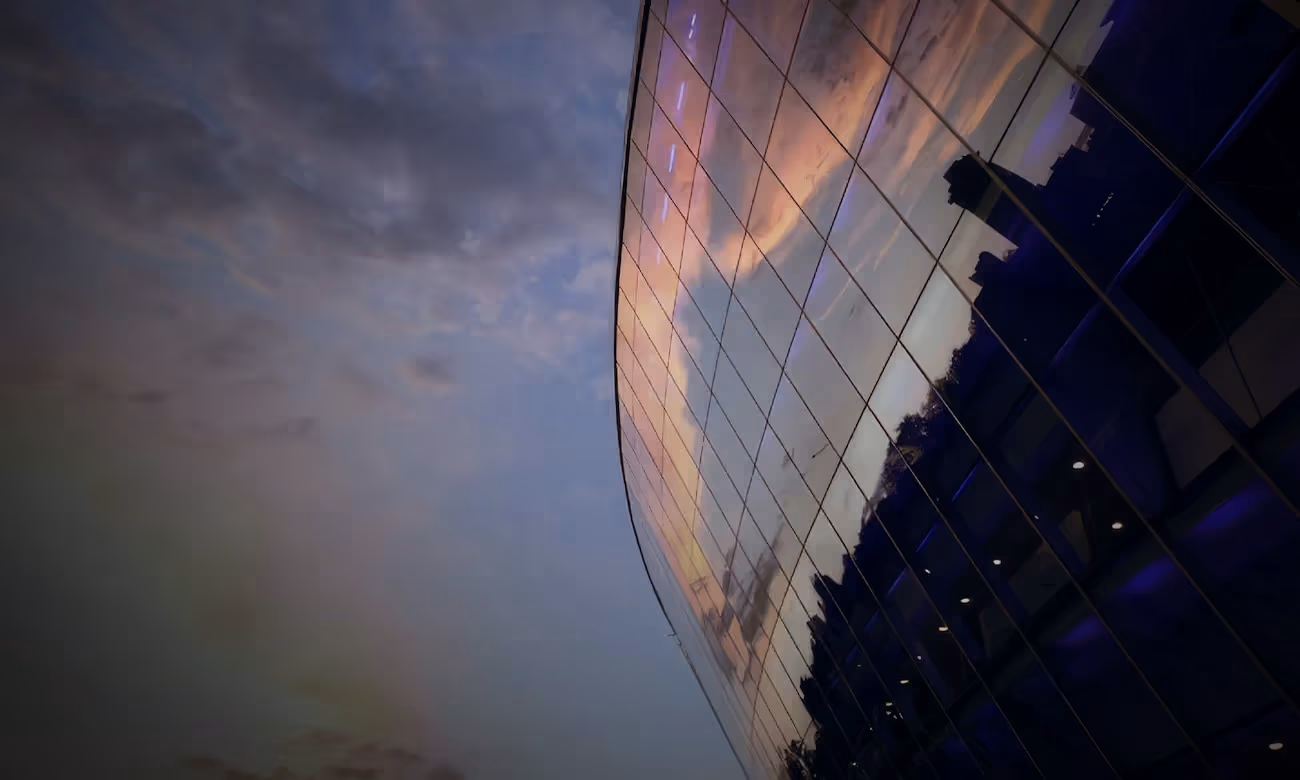 Curved glass building reflecting a purple and orange sunset sky with clouds and city silhouette.