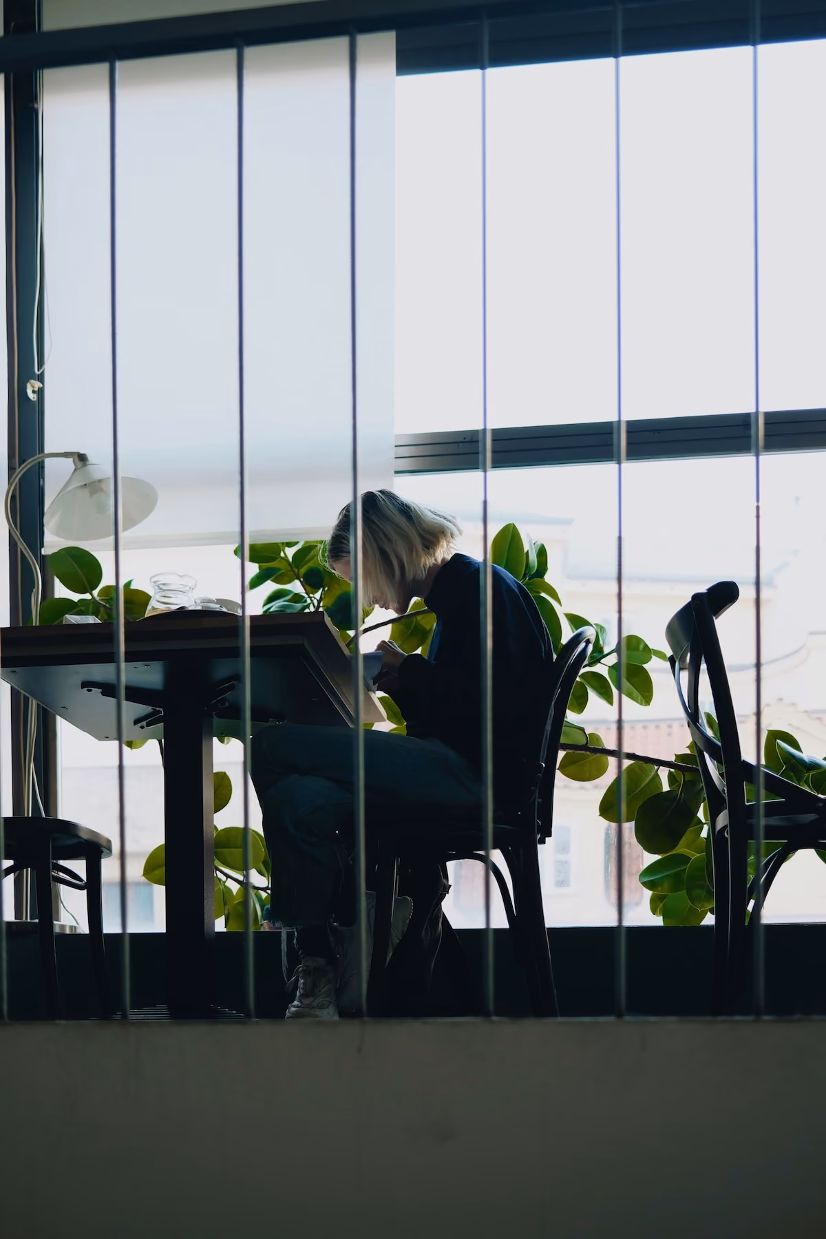 Woman writing at a desk seen through window blinds