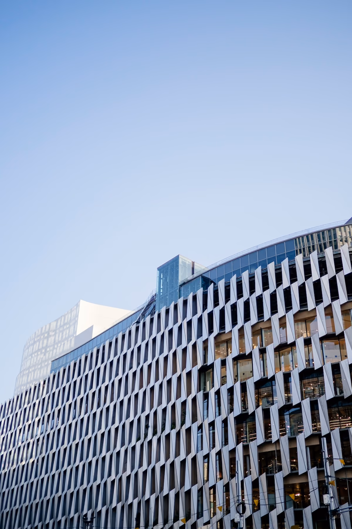 Modern office building with a geometric patterned facade of angled white panels and large glass windows against a clear blue sky.