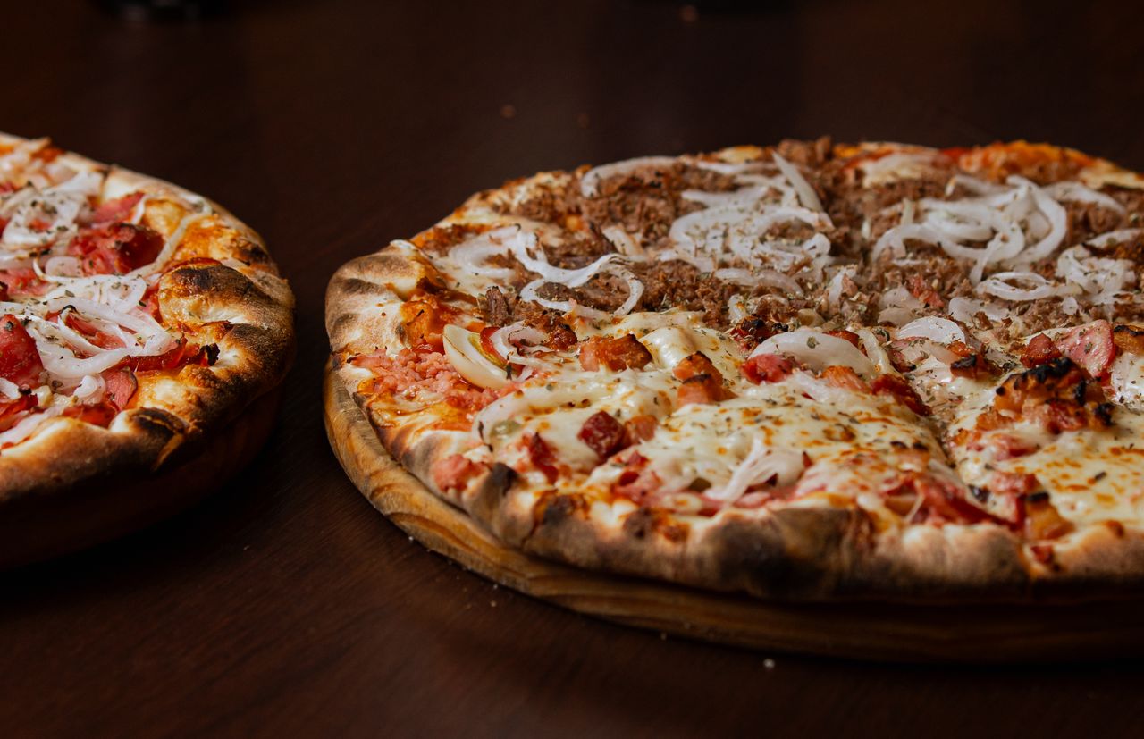 Two pizzas with cheese, onion, and meat toppings on wooden pizza peels against dark background.
