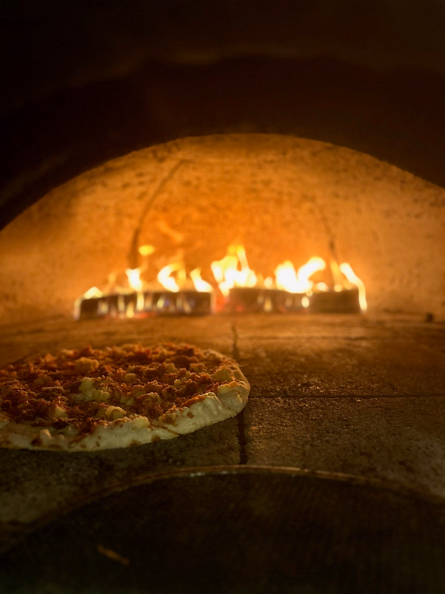 Pizza cooking inside a wood-fired brick oven with flames in the background.