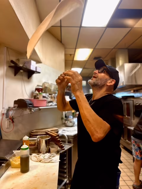 Man tossing pizza dough in a kitchen with shelves, bottles, and ovens in the background.