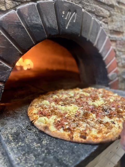 Freshly baked pizza with cheese and meat coming out of a stone oven with visible flames inside.