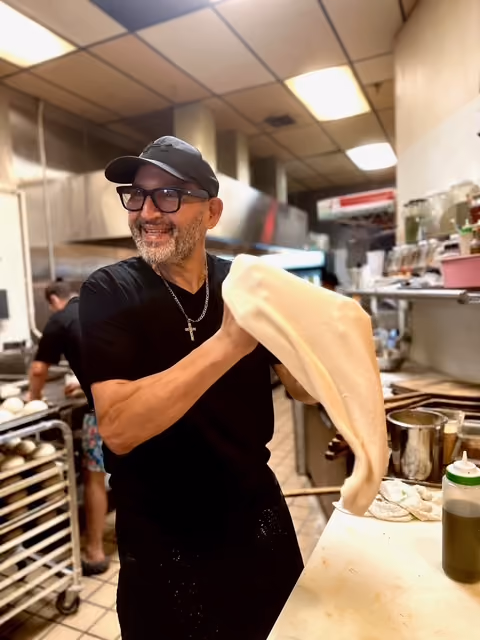 Smiling man tossing pizza dough in a kitchen with pizza dough on a rack in the background.