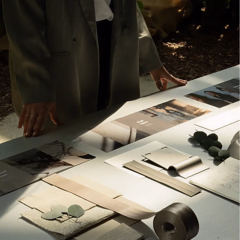 Person in blazer viewing fabric samples, sketches, and print materials laid out on a table in sunlight.