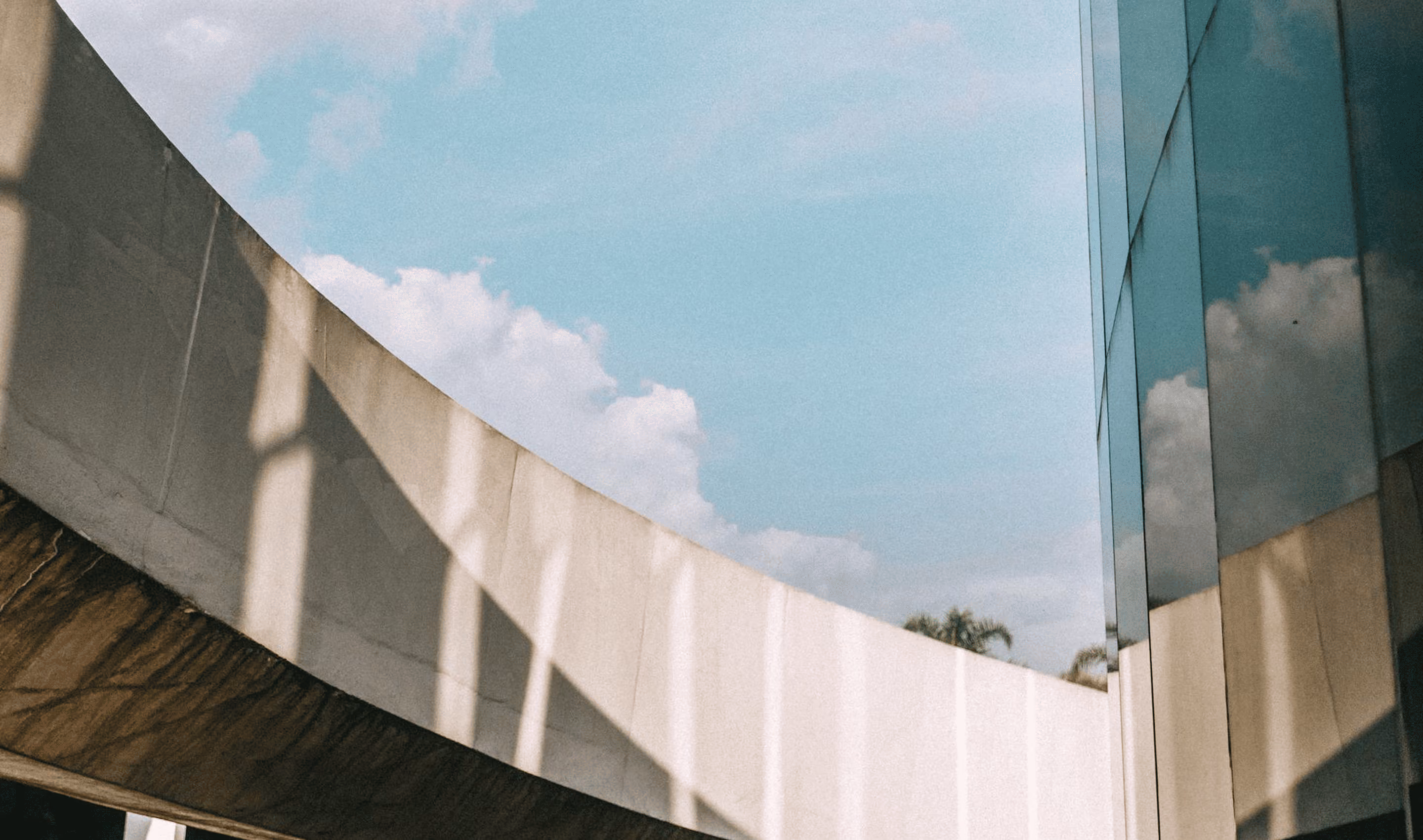 Curved concrete structure with shadows beside a glass building reflecting sky and clouds.