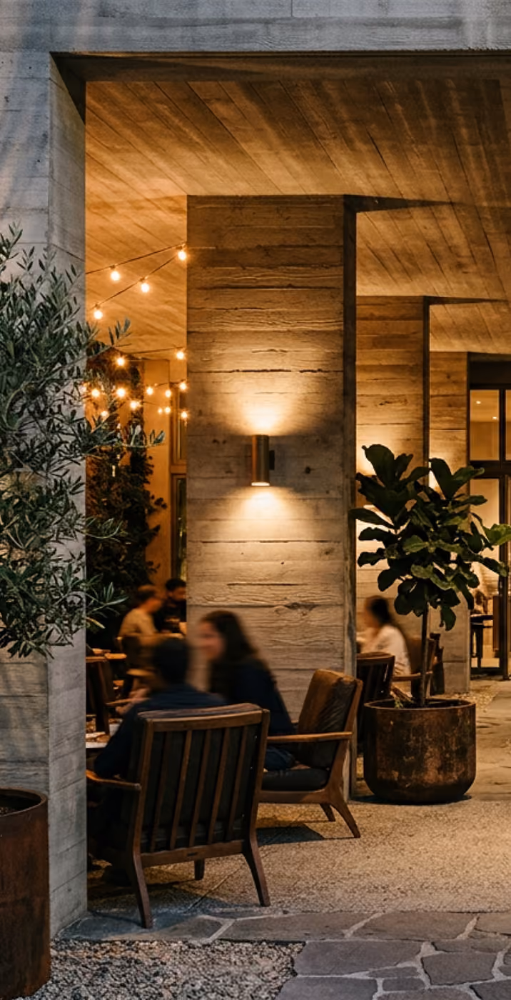Outdoor seating area with string lights, potted plants, and people sitting on wooden chairs under a concrete ceiling.