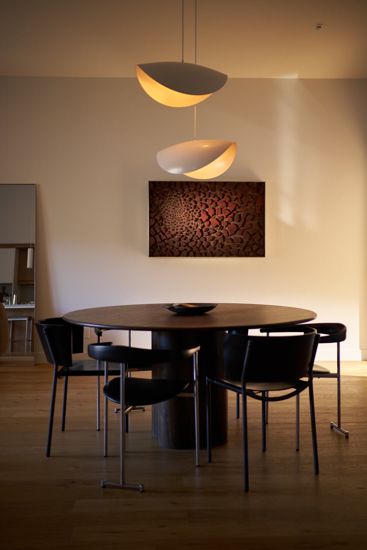 Dining area with round wood table, black chairs, sculptural pendant lights and a textured artwork by Edith