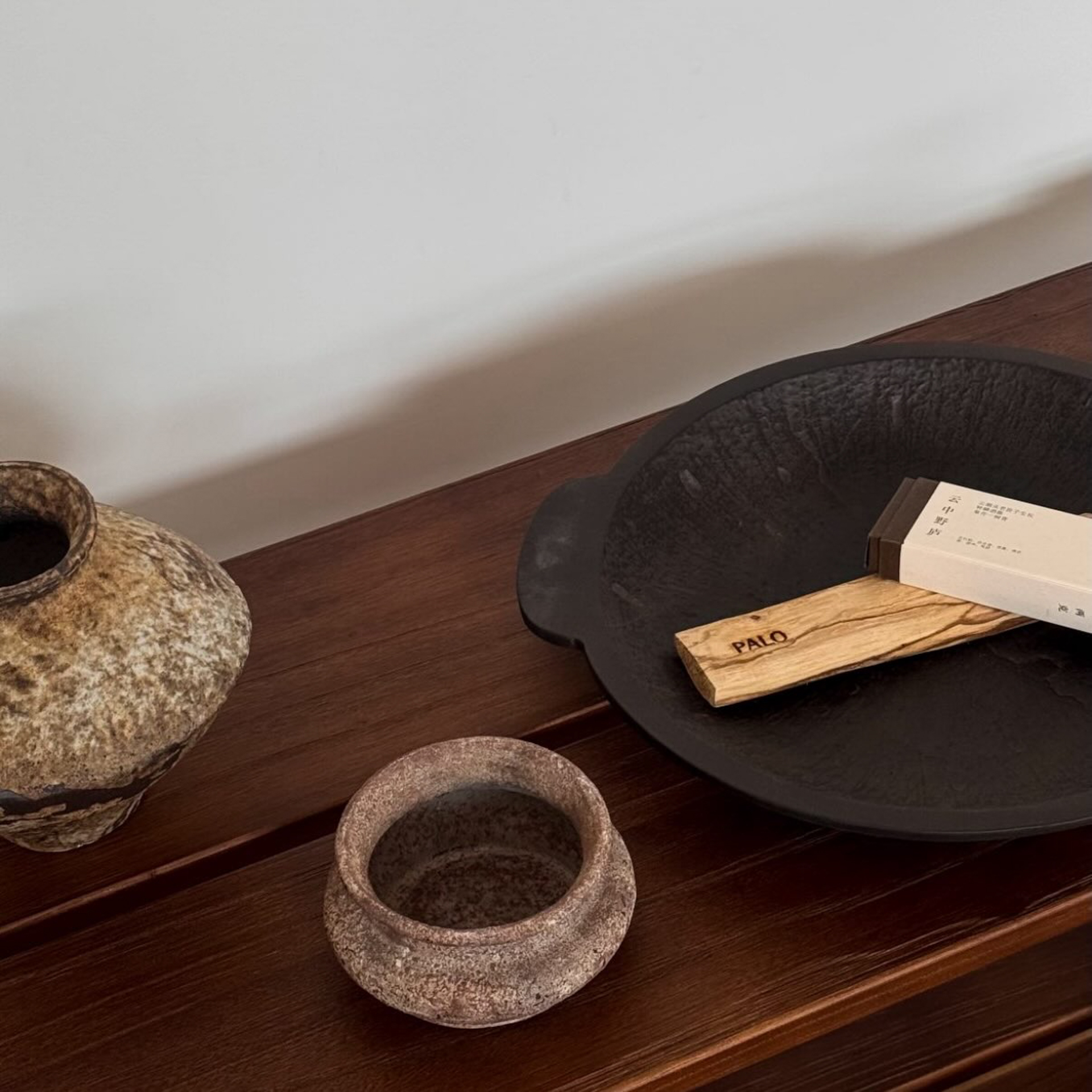 Palo palo santo stick on a dark ceramic dish beside handmade stoneware vessels on a wood console