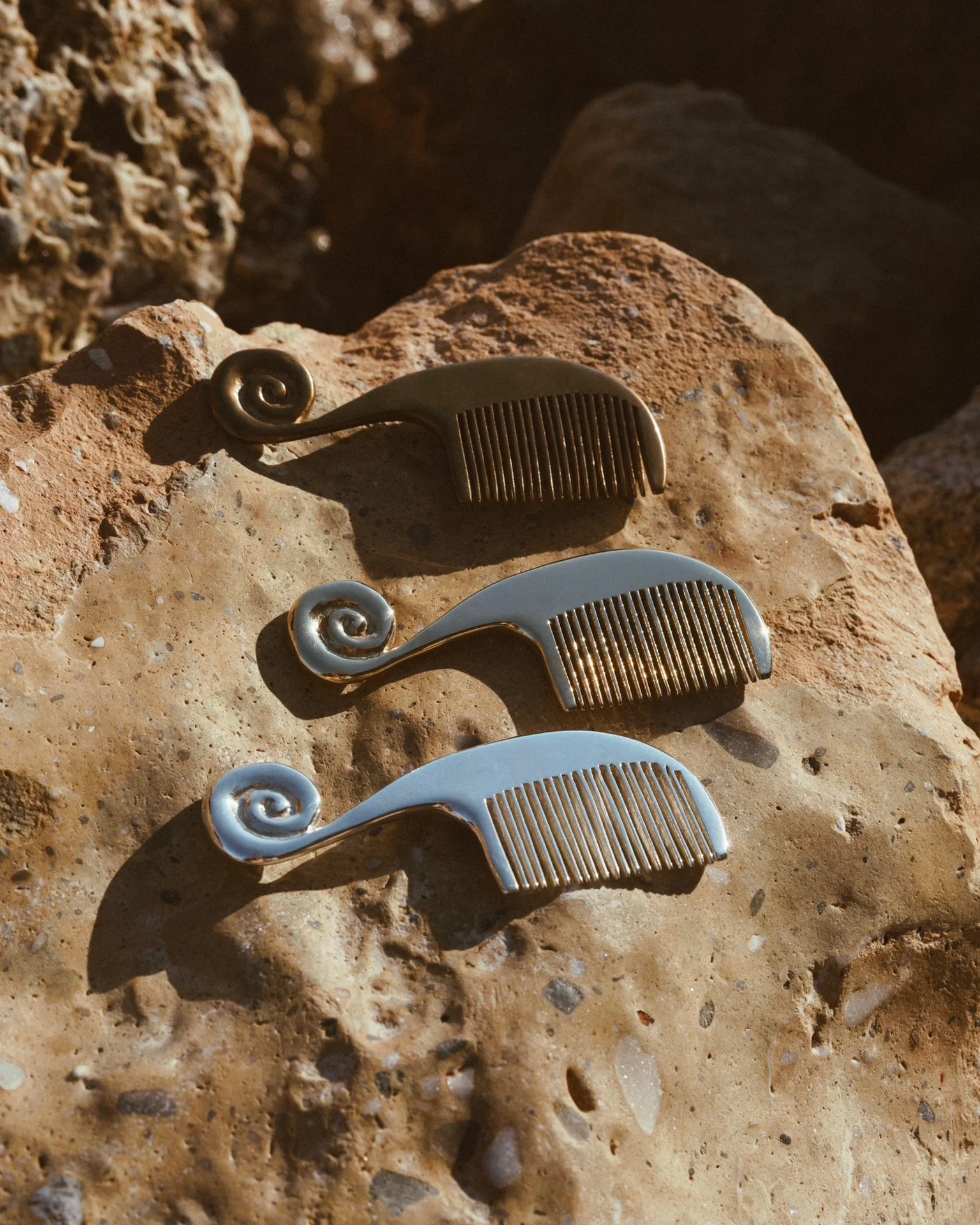 Three spiral-handled metal combs by Jeremy Le Chatelier on sunlit rock, featured in the Rive Lac Tremblant showroom