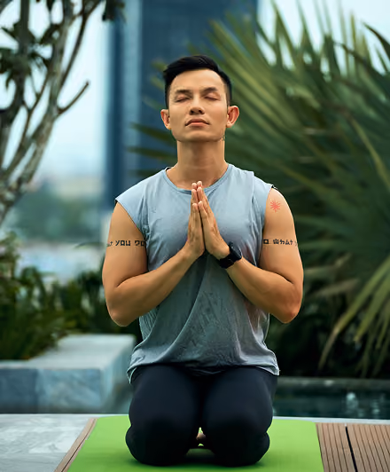 Person with closed eyes kneeling on a green yoga mat with palms pressed together in prayer pose, outdoors near plants and water.