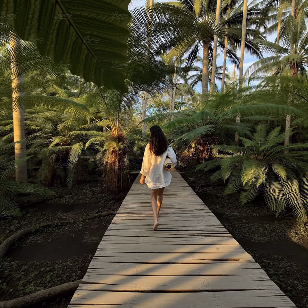 A barefoot woman in a white loose shirt walking on a wooden path surrounded by lush green ferns and tall palm trees in a tropical forest.