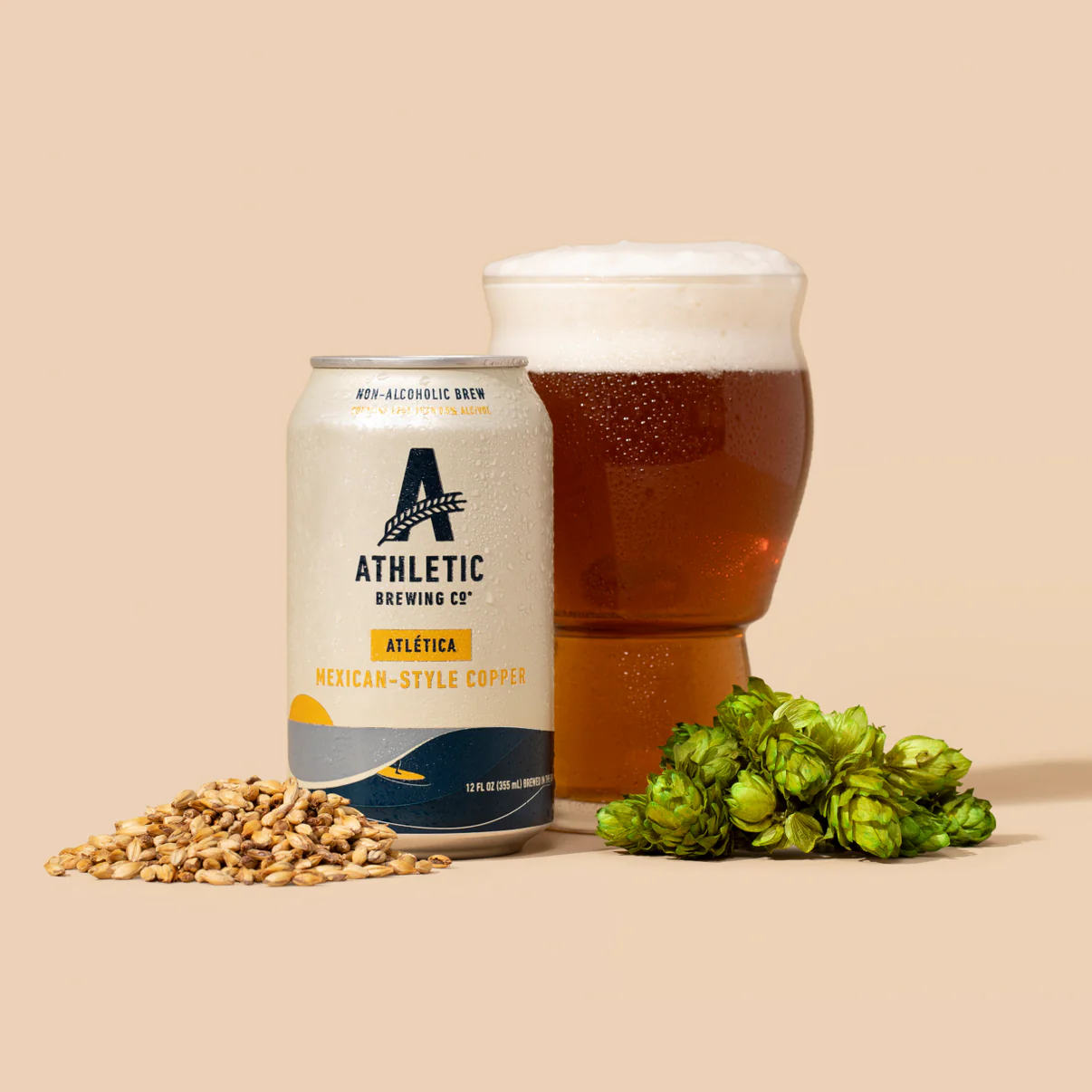 Can of Athletic Brewing Co. Atlética non-alcoholic Mexican-style copper beer beside a full glass, hops, and malted barley