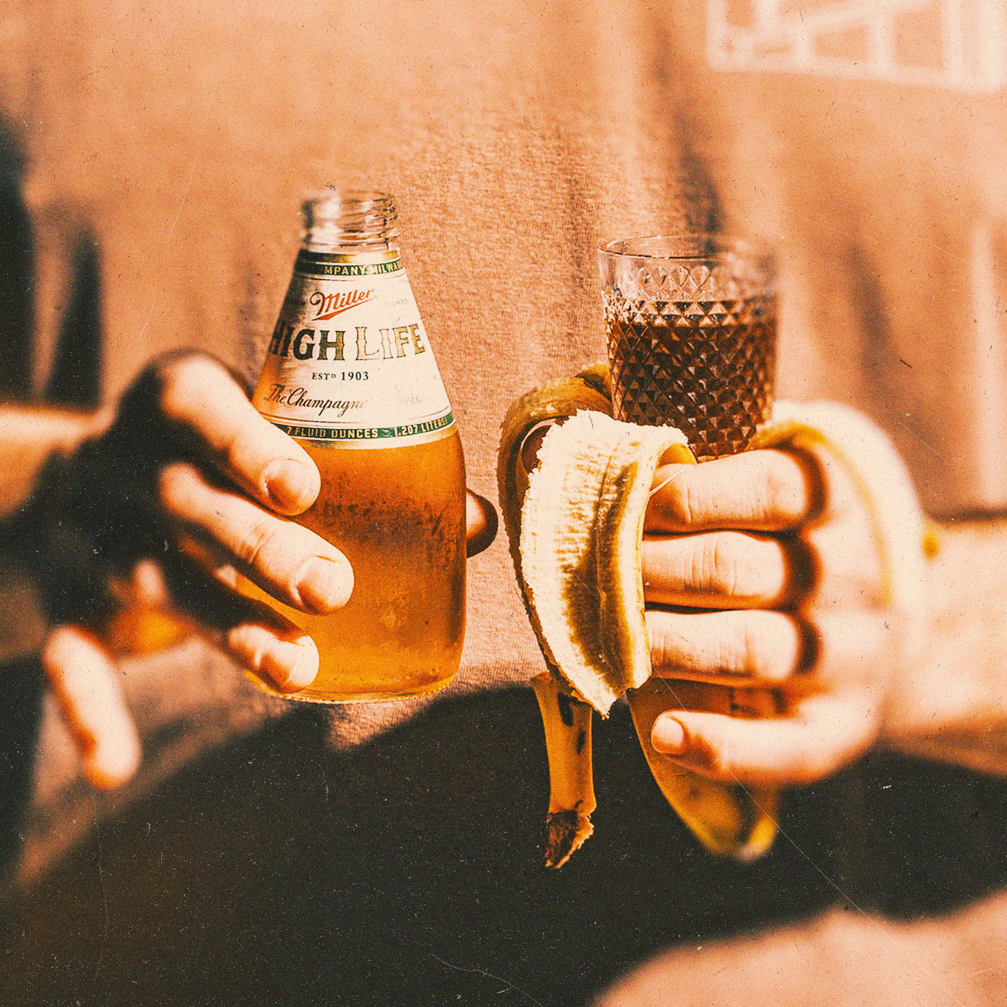Hands holding a Miller High Life pony bottle, a shot glass, and a peeled banana for the Banana Jager Boilermaker