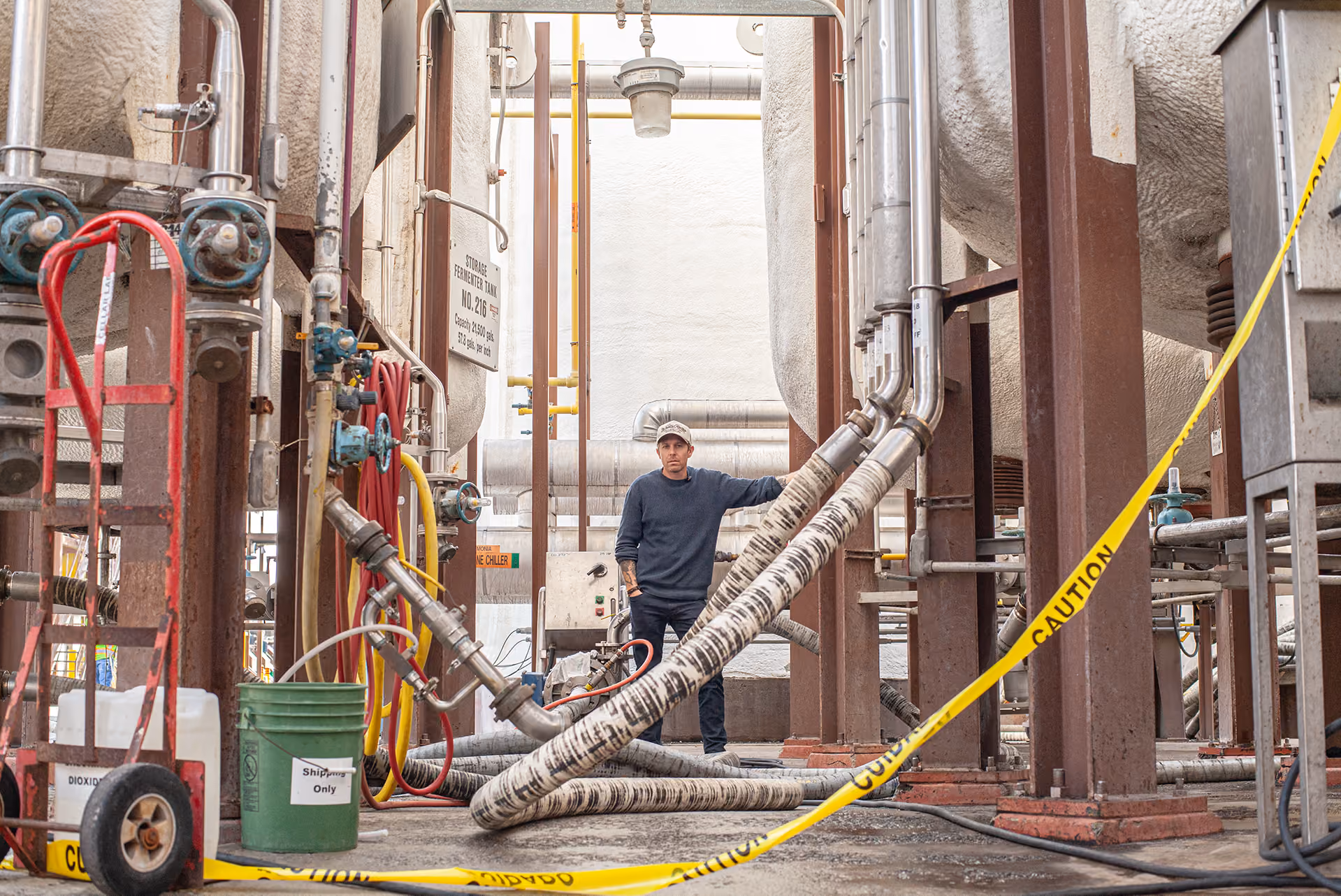 Man in a cap stands among pipes and industrial equipment behind yellow caution tape.