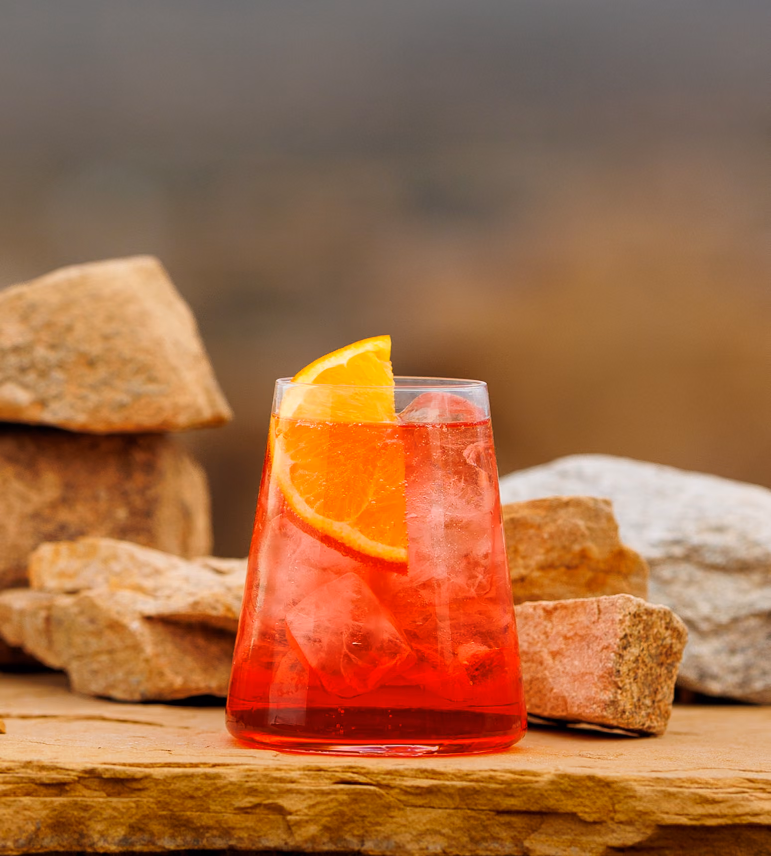 Glass with red drink, ice cubes, and orange slice on a stone surface with rocks in background.