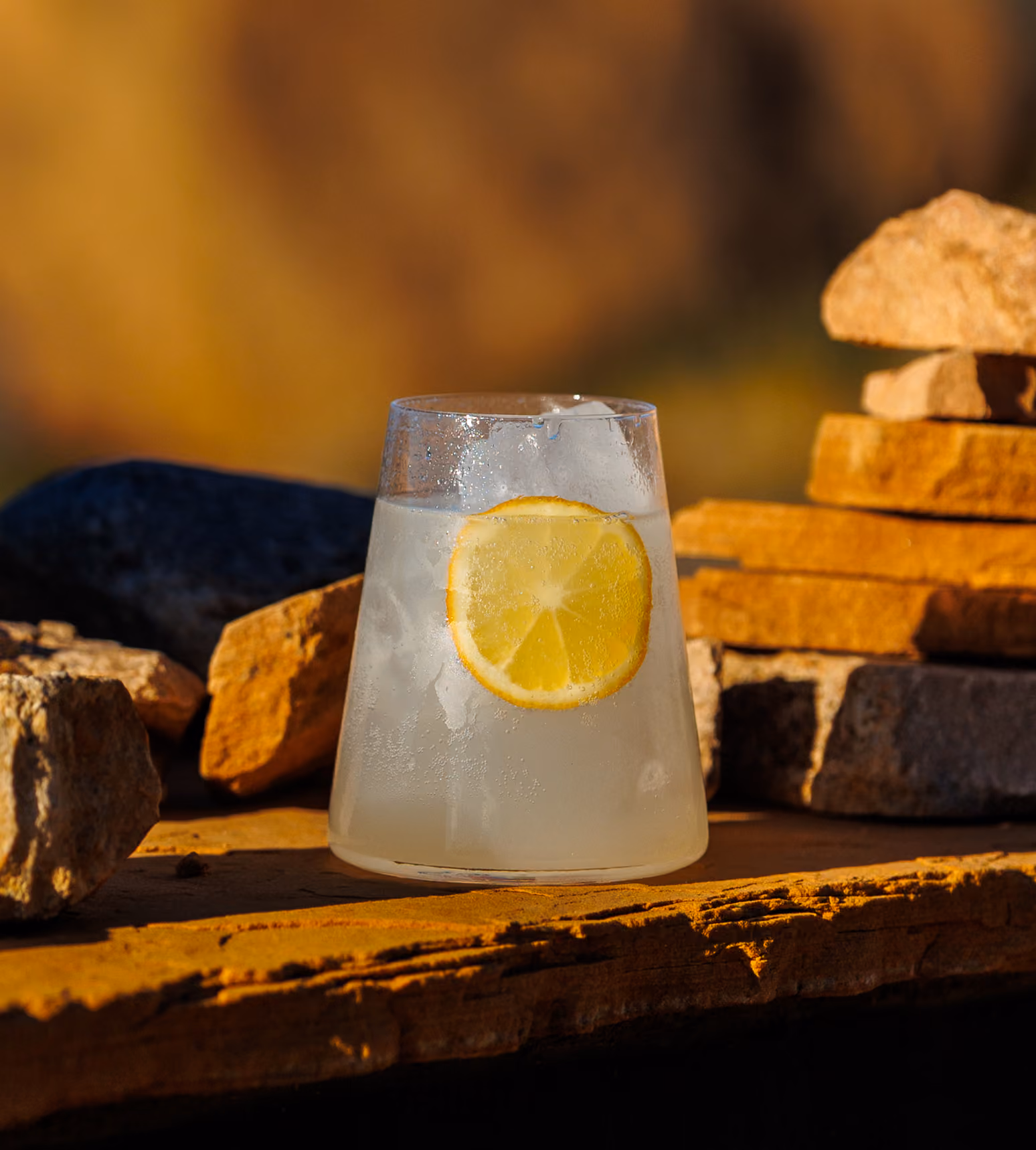 Glass of ice water with a lemon slice resting on a stone surface with rocks in the background.