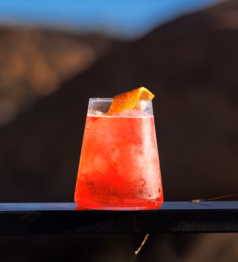 Cold red cocktail with ice and orange peel garnish in a clear glass on a dark surface.
