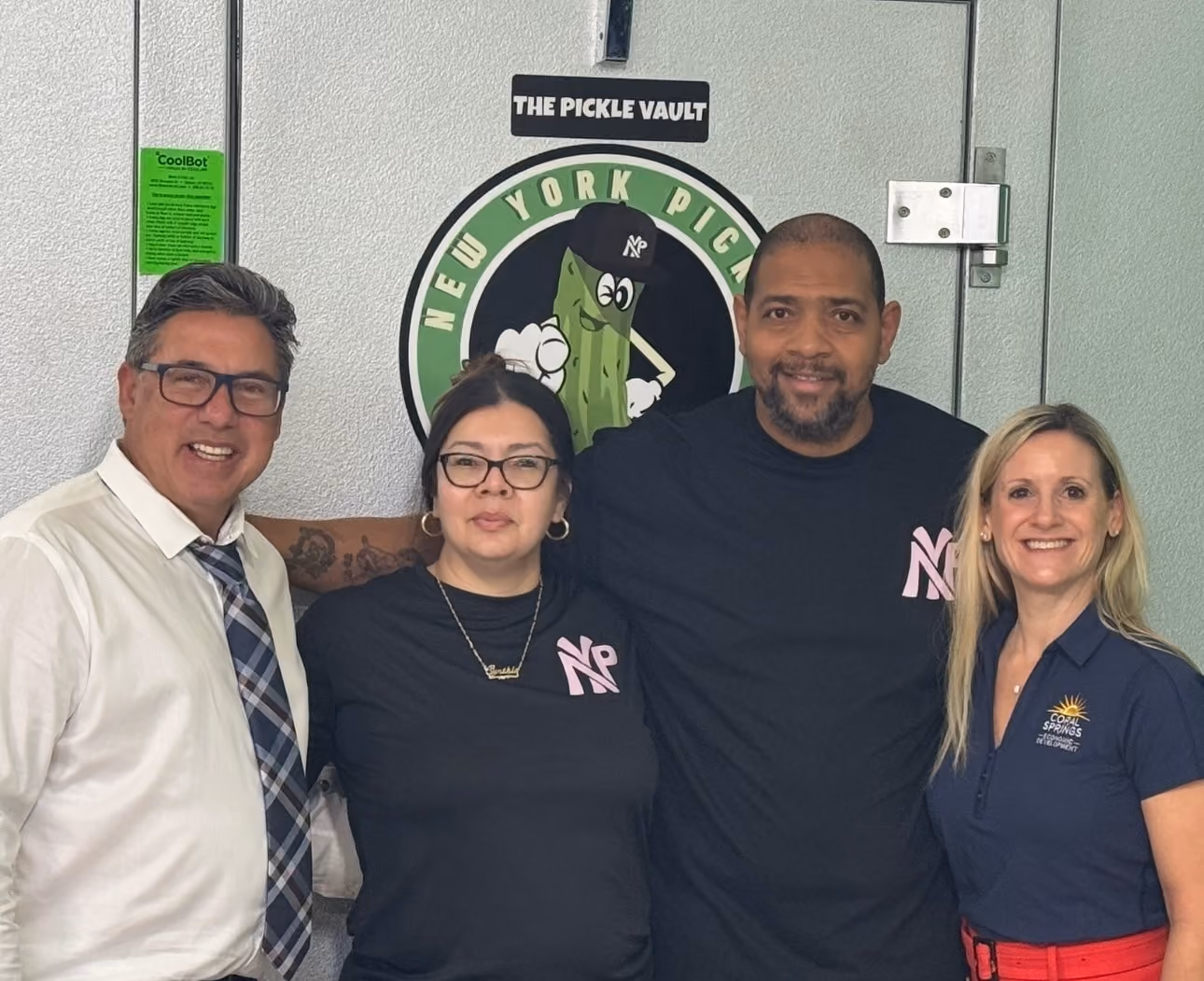 Four people standing together smiling in front of a door with a New York Pickle logo.