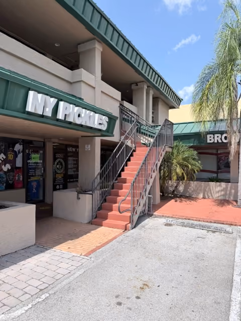 Outdoor view of a building with stairs, green roof, palm tree, and stores with signs including NY Pickles.