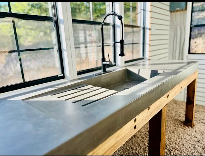 Hand-cast concrete countertop with an integral sink and drainboard grooves, set on a timber base in a window-lined sunroom
