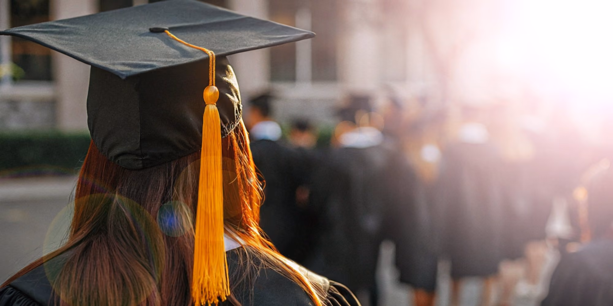 Graduate in cap and gown faces a sunlit commencement crowd outdoors.